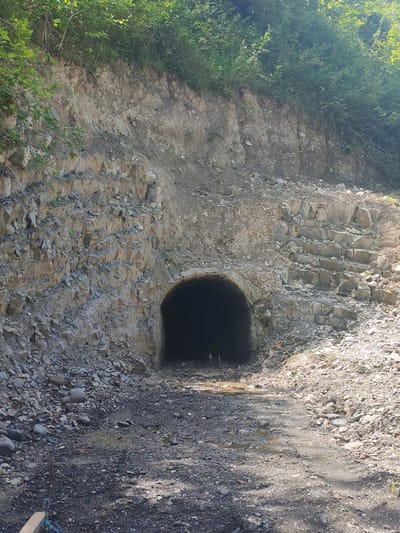 This media file depicts the entrance to a tunnel carved into a rocky hillside, likely in a rural or mountainous area, specifically Orbeli, Georgia. The tunnel opening is dark and appears to be somewhat weathered, with rough-hewn stone surrounding it. The hillside above and to the sides of the tunnel is covered in a mix of loose rocks, dirt, and vegetation, with dense green foliage crowning the top.
The ground leading up to the tunnel is a rough path of gravel and rocks, with some puddles of water suggesting recent rain or a damp environment. The lighting suggests it is daytime, likely late morning or afternoon, with sunlight casting shadows on the ground, particularly on the right side of the path. The weather appears to be fair, as evidenced by the clear sunlight.
There are no people or specific objects visible within the tunnel entrance. However, in the foreground, a small piece of wood and some blue rope are visible on the lower left, indicating some human activity or presence in the vicinity, possibly related to construction, maintenance, or exploration of the tunnel. The overall scene evokes a sense of rugged natural beauty and perhaps a hint of mystery due to the dark, unknown interior of the tunnel. The layered rock formations on the right side of the tunnel entrance are a notable geological detail. No visible text is present in the image.