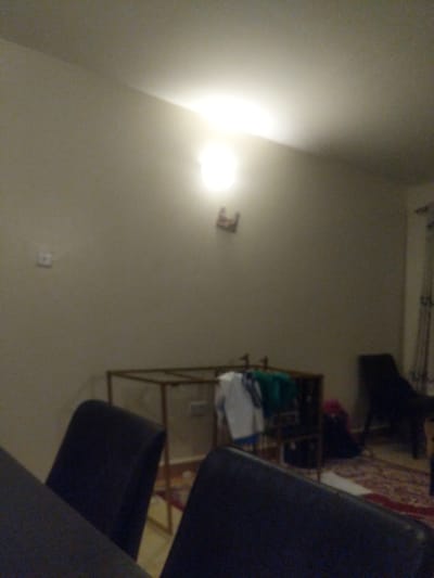 The image appears to be taken indoors in a residential setting in the Kitengela ward of Kenya. The scene is dimly lit by an overhead light fixture. The walls are painted a light beige color. A partially visible metal shelving unit contains folded clothing items, including a green shirt and white items. A chair is in the foreground, obscuring part of the scene. The room is cluttered, with an area rug visible in the background. Other items are present but indistinct. There is no activity of people. A wall switch and a fire alarm device are affixed to the wall.