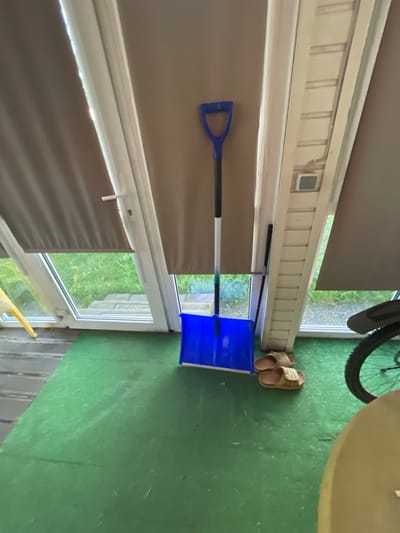 In what appears to be a sunroom or enclosed porch area in Bol'shoe Zaborod'e, Russia, a bright blue snow shovel leans against a wall near a set of sliding glass doors. The doors are covered by beige pull-down blinds, with a sliver of the outside greenery visible through the windows. On the green floor, a pair of tan flip-flops are placed next to the shovel. To the right, the rear wheel and part of the frame of a bicycle are visible, suggesting it is parked nearby. The overall impression is one of a functional, possibly utilitarian, space, perhaps preparing for or storing winter equipment.