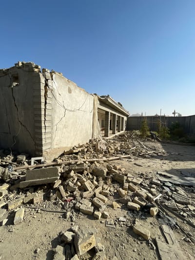 The image displays a scene of significant structural damage to a single-story building in Spin Boldak, Afghanistan, under a clear, bright blue sky. The light grey concrete structure exhibits prominent vertical and diagonal cracks along its exterior walls, particularly at the left corner. The upper portion of the building's facade is broken, with sections of the roofline compromised or removed. A white sheet or tarp hangs from the wall in the central portion of the building. Further to the right, a section of the building features a covered veranda supported by a series of grey pillars.
The ground in front of and around the building is covered with extensive rubble. This debris primarily consists of fragmented light-beige bricks, many stamped with the word "PAKISTAN," mixed with broken concrete pieces, wooden planks, and other construction materials. The terrain is unpaved, dusty earth.
In the background, a tall concrete perimeter wall runs horizontally, behind which sparse vegetation and trees are visible. A black motorcycle is parked near the wall, partially obscured. Distant, slender, dark cylindrical structures, possibly minarets or communication towers, are discernible against the hazy horizon. No people are present, and no actions or interactions are depicted in the static scene. The environment is arid and dry.