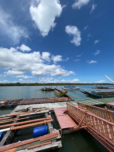In the city of Ko Panyi, Thailand, a waterfront scene unfolds under a bright blue sky dotted with fluffy white clouds. Several wooden boats are docked, their colorful hulls resting on the water. A floating wooden dock, partially constructed with unfinished wood planks and blue plastic barrels, extends into the calm water. Another section of the dock features a red wooden walkway with an ornate gold railing, leading to the boats. In the distance, a lush green treeline borders the water, with a few islands visible on the horizon. A white pole stands tall on the right side of the frame.
