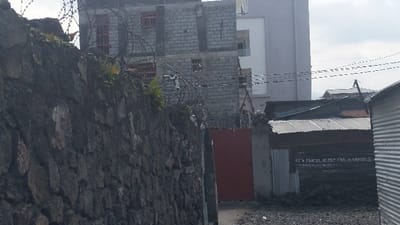 The image shows a narrow alleyway in Rubavu, Rwanda, located at coordinates -1.668 latitude, 29.268 longitude. The alley is bordered on the left by a tall stone wall and on the right by a building with a corrugated metal roof and the words "Cete Parcel N'est Pas a Vendre" (This Parcel is Not For Sale) written in white paint. The alley leads to a red metal gate in the foreground. The buildings are constructed of concrete block and appear to be under construction. The alley is covered with a layer of loose gravel and there are no people present.