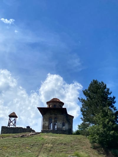 The image shows a historical complex located on a grassy hilltop under a bright blue sky with scattered white clouds. The complex includes ancient stone structures and a prominent, partially ruined church with a red tiled dome. To the left of the church, there is a wooden bell tower with a similar red tiled roof, situated on a stone foundation. The foreground is dominated by a grassy slope with some sparse vegetation. A large evergreen tree stands to the right of the church. The scene appears to depict an archaeological site or a historical monument.

The location is the Gradina archaeological site, also known as the Church of St. Peter and Paul, situated in the municipality of Raska, Serbia. This site represents the medieval capital of the Serbian state, Ras, which flourished between the 8th and 12th centuries. The complex features remnants of fortifications, churches, and other structures dating back to that period.