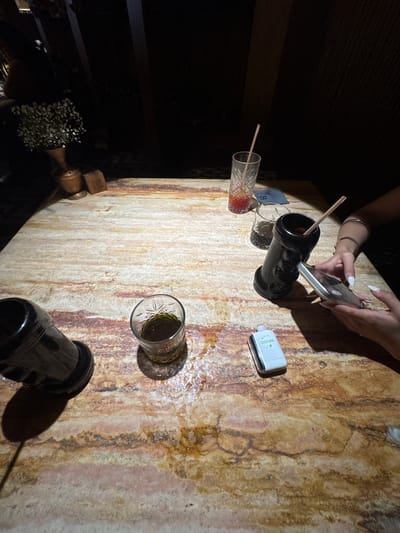 Captured at an overhead angle, the image reveals a table scene likely situated in San Pedro Garza García, Mexico. The tabletop, displaying a distinctive marbled texture, is populated with several items. A person's hands, adorned with bracelets and manicured nails, hold a smartphone. Next to them, a black, uniquely shaped glass contains a straw. Adjacent to this is a white Geek Bar disposable vape device. Further down the table, a cylindrical glass, partially filled, rests near a similar black glass. Another drinking glass, also containing liquid and a straw, lies on its side. A small bouquet of white flowers, along with other indiscernible items, sits towards the edge of the table. The lighting is dim, contributing to a slightly shadowed ambience.