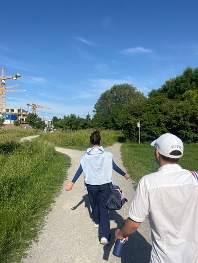 The image shows two people walking along a gravel path that forks into two directions. The path is surrounded by green grass and trees, and there is a construction site in the background. The woman in the foreground is wearing a light blue shirt, dark blue pants, and a blue backpack. She is walking to the right side of the image, away from the camera. The man is walking to the left of the image, towards the camera. He is wearing a white shirt, a white cap with the words "ROY CONRAD" written on it and red and white striped fabric around his wrist. He is holding a blue water bottle in his right hand. The scene is located in Vienna, Austria, at the coordinates 48.246, 16.423.