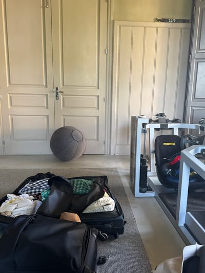 The image shows a light gray door with silver hardware, positioned beside a dark gray, round pouf resembling the Death Star. Adjacent to the pouf are two open black suitcases with clothing items inside. A silver exercise machine stands above the suitcases on a dark gray carpet. A decorative ribbon is seen above the door, possibly indicating a year.