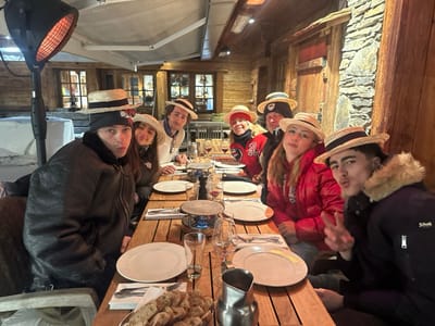 The image depicts seven individuals, primarily young adults, seated at an outdoor wooden table during nighttime or dusk in La Clusaz, France. The setting is a rustic restaurant terrace, characterized by dark wooden walls, exposed stone masonry, and illuminated windows in the background. An overhead outdoor heater with a visible glowing orange element is positioned on the left.

On the table, there are multiple white dinner plates, wine glasses, cutlery, a metal fondue pot heated by a blue flame, a basket of sliced bread, and a metal pitcher.

Of the seven individuals, four are wearing straw boater hats. A male on the far left, wearing a dark leather jacket with a fleece collar, is posing with pursed lips. To his right, a female with dark hair is partially visible. Next to her, a male in a white sweater is seated. Across the table, from left to right, are a female in a red puffer jacket with an "A" logo, a male in a dark beanie and dark puffer jacket, a female in a red puffer jacket, and a male on the far right in a dark, fur-lined puffer jacket (Schott brand visible). The male on the far right is making a peace sign gesture with his right hand and has pursed lips. Snow is visible on a surface to the far left of the dining area.