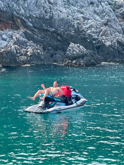 In Dubraljevina, Montenegro, a man is relaxing on a jet ski in clear turquoise water near a rocky coastline. The man, wearing checkered shorts, is leaning back on the jet ski with his legs extended and his left arm raised. A red bag is secured behind him on the jet ski. The water is slightly rippled, with sunlight glinting off the surface. The rocky shore, characterized by its grey, textured surface, forms the backdrop to the scene. The jet ski has "YAMAHA" branding visible on its side.