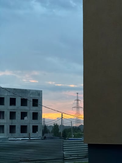The image, taken in the city of Froly, Russia, depicts a construction site during dusk. In the foreground, a large, unfinished concrete building with square openings for windows is visible on the left. A corrugated metal barrier runs horizontally across the bottom of the frame. To the right, the textured, beige wall of another structure dominates the right side of the image, obscuring a portion of the scene. In the background, a tall, lattice-like electrical pylon stands against a sky painted with the soft hues of sunset, featuring streaks of orange and pink clouds against a pale blue. Trees and other distant structures are silhouetted against the horizon. Power lines are strung across the mid-ground. No people are present in the image.