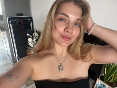 A woman is taking a selfie. The image appears to be taken indoors. She is wearing a black top and a necklace with a decorative pendant. She is wearing beaded bracelets on her left wrist. A plant and a framed picture are visible in the background. A white wall and part of what appears to be a kitchen appliance can be seen. There is no information to indicate that the photograph was taken in Большая Шильна, Russia.