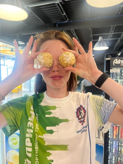 The image shows a woman holding two wrapped rice crispy treat balls in front of her eyes, as if they are her eyes. She is indoors, likely in a shop or store. She is wearing a white t-shirt with green and blue accents, featuring an emblem with the text "SPORT TOUR RUSSU" and a design that resembles a cityscape with churches. The shirt also has Cyrillic text that appears to be related to travel or prizes. Her left wrist has a black activity tracker, and there's a ring on one of her fingers. The background has overhead lighting and a display of products, possibly snacks or drinks. Based on the church silhouette on the shirt, it is potentially in Казань, Russia.