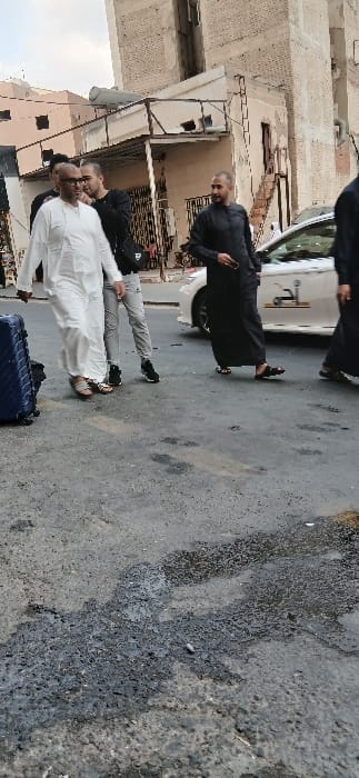 The image depicts an urban street scene in Makkah Al Mukarramah, Saudi Arabia, under an overcast sky. In the foreground-left, a bald man with dark skin and glasses, wearing a white thobe and brown sandals, walks rightward, pulling a blue rolling suitcase, partially visible in the lower-left corner. Immediately behind him and slightly to his right, a younger man with a beard, wearing a black long-sleeved shirt, light grey trousers, and black athletic shoes, walks in the same direction, carrying a black shoulder bag. To their right, another man with a beard, dressed in a black thobe and black sandals, also walks rightward, holding a dark, elongated object in his right hand. A fourth individual in a black thobe is partially visible on the far right. A white sedan, possibly a taxi or ride-hailing vehicle with a dark Arabic decal on its side, is parked or moving slowly on the right side of the street. The asphalt road surface shows significant wear, with cracks, patches, and several dark, wet-looking stains. In the background, multi-story residential or commercial buildings with aged, light-colored facades, exposed brickwork, and visible utility structures such as a satellite dish and external staircases are prominent.