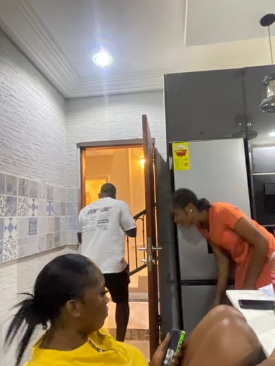 This indoor photograph, taken in Adenta Municipality, Ghana, captures a casual scene involving three individuals within a home environment. The image is rotated 90 degrees clockwise.
In the center, a woman dressed in an orange top is bending forward, appearing to interact with or look into the lower section of a dark grey or black refrigerator. A yellow energy efficiency label, featuring red text and graphics, is prominently displayed on the refrigerator door. To the left, a bright, circular ceiling light fixture emits a strong glow, illuminating the space.
In the middle ground, a man wearing a white t-shirt and dark bottoms stands with his back to the camera, positioned within a wooden-framed doorway or window opening that leads into another brightly lit room. The walls throughout the visible space exhibit a textured finish.
In the foreground, on the right, a second woman in a yellow top is partially visible, holding a mobile phone or similar device in her hand. The floor includes a section of patterned tiles featuring blue and white geometric designs, alongside a lighter, textured floor area. The atmosphere appears relaxed and domestic. No specific emotions are clearly discernible from the visible faces. Text on the man's white t-shirt is present but illegible.