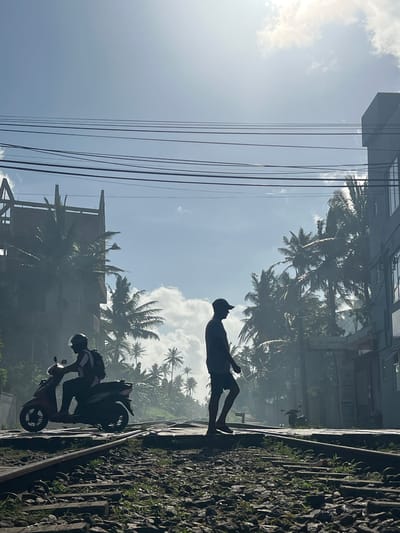 The image depicts a silhouette of a person standing on a railway track, facing away from the camera and looking towards a distant building. The person is wearing a hat and appears to be casually dressed. In the foreground, a red scooter with a person on it, also silhouetted, is positioned on the tracks. The scene is set amidst lush green palm trees and a bright, cloudy sky, suggesting a tropical or subtropical environment. The railway tracks themselves are covered with gravel and interspersed with small plants. Overhead, numerous power lines crisscross the sky. In the background, modern buildings are visible, juxtaposed with the natural elements and the rustic railway. The lighting suggests it is daytime, likely late afternoon given the long shadows and the warm glow of the sun. The overall mood is peaceful and serene, with a hint of everyday life unfolding in a picturesque setting. There is no visible text in the image. The location is identified as Kodagoda, Sri Lanka.