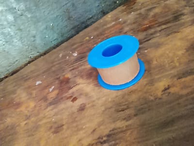 A roll of opaque, beige tape with a bright blue plastic core is positioned on a rough, weathered wooden surface. The wood exhibits a grain pattern with variations in color, including shades of brown and reddish-orange. Some white specks, possibly dust or debris, are scattered across the wood. In the background, a textured, pale blue or gray surface with an uneven, speckled appearance is visible, creating a contrasting backdrop to the wooden foreground. The lighting suggests it might be daytime, with the light source casting subtle shadows. There is no visible text in the image.