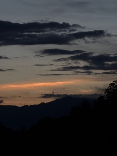 This is a landscape photograph of a dramatic sky at dusk, likely taken in Choumkhong, Laos. The image is oriented vertically. The foreground is dominated by dark silhouettes of trees or foliage on the right side, creating a stark contrast with the sky. The sky itself is a spectacle of layered clouds. On the left, there are heavy, dark grey clouds with textured, streaky formations, suggesting a brooding or overcast condition. Moving towards the center and right, the clouds become lighter and are illuminated by the setting sun, displaying hues of orange, yellow, and soft peach. These brighter clouds are also streaky but appear more ethereal and wispy. The transition from dark to light in the sky is striking, hinting at the moment between daylight and night. There is no discernible text in the image. The overall mood conveyed is one of natural beauty and the transition of time, potentially evoking feelings of awe or tranquility.