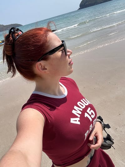 A red-haired woman wearing sunglasses and a red t-shirt with white lettering that reads "Amour 15" is lying on a sandy beach next to the ocean. The ocean waves are visible in the background. The woman is fair-skinned and has freckles. A backpack is visible near her arm. She is looking towards the ocean with a relaxed expression.