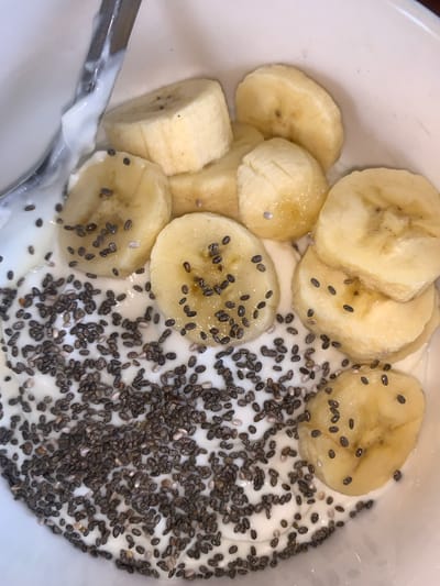 This image depicts a bowl of yogurt topped with sliced bananas and chia seeds. The yogurt is white and creamy, and the chia seeds are small, black, and densely scattered across the yogurt. The banana slices are arranged on top of the yogurt, with some chia seeds also adhering to them. A spoon is visible at the bottom left edge of the bowl, partially covered in yogurt. The scene appears to be a close-up shot of a food preparation, possibly a breakfast or snack. The image does not show any location or other elements to infer that this image was taken in Galadimawa, Nigeria.