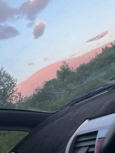 The image shows a view from inside a vehicle, looking out through the windshield and side window at a sky filled with pink and blue clouds. The clouds are illuminated by the setting sun, creating a vibrant sunset effect. Below the sky, there is a landscape of green trees and vegetation, with a dirt road or path visible in the distance. The foreground of the image is dominated by the interior of the car, including the dashboard, windshield wipers, and a portion of the steering wheel. The overall impression is one of a scenic drive during a beautiful sunset. This image was taken in Sovetskaya Gavan', Russia.