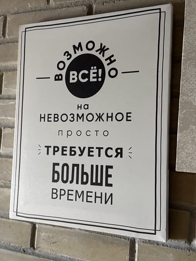 The image shows a sign in Russian, which translates to "Everything is possible! It just takes more time." The sign is white with black lettering and a black border. It appears to be hanging on a brick wall. The location in the city of Zamankul, Russia, is not discernible from the image.