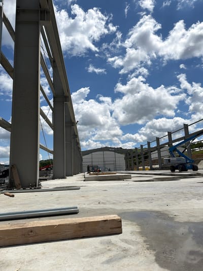 This is a daytime, low-angle, outdoor photograph of a construction site. The foreground is a concrete surface with some puddles and scattered debris, including a wooden beam and a metal conduit. In the mid-ground, a row of large, grey steel I-beams form the skeletal structure of a building. Beyond these beams, more steel framework for a larger structure is visible, along with a white building with a curved roof. A blue Genie boom lift is positioned in front of the white building. The background features a treed hillside and a bright blue sky filled with fluffy white clouds. The scene suggests ongoing construction activities in Skrzydlna, Poland. There are no individuals clearly visible in the image.