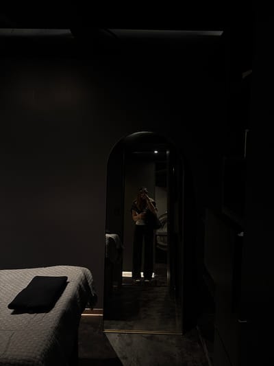 The image depicts a dark room with a large, arched mirror reflecting a person taking a selfie in what appears to be a bathroom or dressing area. The reflection shows a person wearing dark clothing and holding a phone or camera. The mirror is framed by a dark surface, possibly a wall or furniture. In the foreground, a bed with a textured white bedspread and a dark square object (possibly a towel or blanket) is visible. The room is dimly lit, with the main light source coming from behind the person reflected in the mirror. Based on the prompt, this scene is located in Ciudad López Mateos, Mexico.