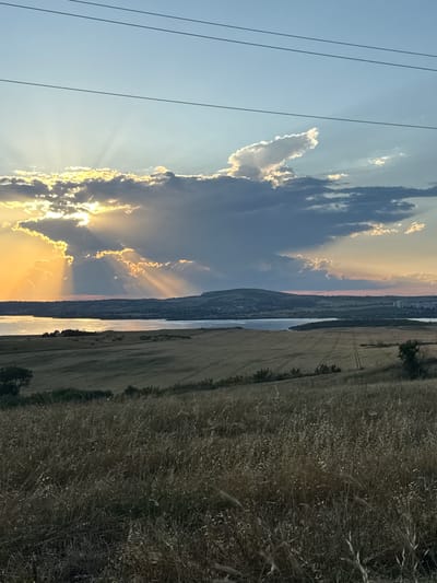 The image captures a panoramic view of a landscape at sunset, featuring a body of water, rolling hills, and cultivated fields. In the foreground, tall, dry grass dominates, casting shadows that suggest the low angle of the sun. Beyond this, a vast field, possibly wheat or barley, shows distinct tire tracks from agricultural machinery, indicating recent harvesting or cultivation. Further in the distance, a wide expanse of water, likely a reservoir or a large river, reflects the warm hues of the setting sun. The horizon is marked by a range of hills, some sparsely populated with vegetation and buildings, with a notable cluster of structures visible on the right side, suggesting a settlement.

The sky is a dramatic canvas of light and shadow. A large, dark cloud formation partially obscures the sun, creating striking crepuscular rays that pierce through the clouds, illuminating the sky with golden light. The sun itself is low on the horizon, casting an orange glow. A few power lines are visible in the upper portion of the image, cutting across the sky.

This scene is located in the vicinity of Tvarditsa, Bulgaria, a region known for its agricultural lands and proximity to water bodies such as the reservoir named after the town. The time of day is late afternoon or early evening, evidenced by the long shadows and the golden light. There are no people or animals visible in the frame, making it a serene and undisturbed landscape composition. The overall mood is one of tranquility and natural beauty, highlighted by the dramatic interplay of light and cloud formations during sunset.