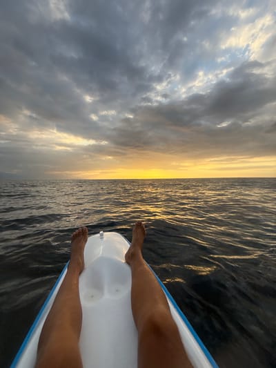 The image shows a person's legs and feet from a first-person perspective, resting on the front of a white and blue boat. The boat is on the water, and the horizon is visible in the distance, marked by a bright yellow sunset. The sky is filled with dramatic, dark clouds, with some lighter patches where the sun's rays are breaking through. The water is dark and rippled, reflecting the colors of the sky. The location is specified as the city of Buleleng, Indonesia.