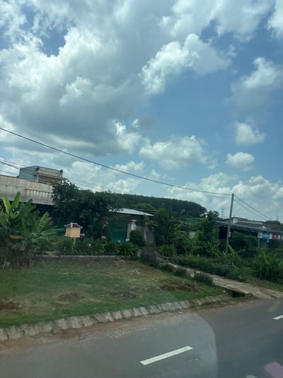 An exterior, eye-level, medium shot captures a roadside view under a cloudy, blue sky. The foreground features an asphalt road with a white dashed line. A concrete curb separates the road from a grassy area with some bare patches and small piles of dirt. Behind the grass, a concrete path slopes upwards, leading to a verdant scene with lush green foliage, including banana trees.
Several structures are visible in the background. On the left, a multi-story building with a light blue upper section and a white facade stands behind a decorative railing. A small, yellow, pagoda-like structure is situated among the plants in front of a green fence. Further back, a long, low building with a green roof and an open-fronted structure with metal supports stretches across the scene. On the right side of the image, more buildings are visible, some with awnings and signs, including ones for "Viettel" and "Intesphone." The background is dominated by a line of trees and distant utility poles with power lines crisscrossing the sky. The overall impression is a semi-rural or suburban landscape.