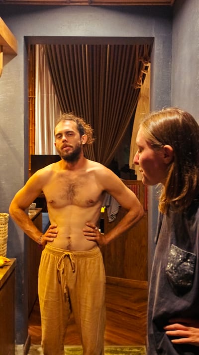 The scene is set indoors in Mu Cang Chai, Vietnam, featuring two individuals illuminated by warm, subdued lighting. A male subject, shirtless and wearing light brown drawstring pants, stands centrally on the left. He presents with a beard and mustache, and his dark, curly hair is gathered at the back of his head. Visible body hair covers his chest and abdomen. He stands with both hands placed on his hips, elbows outward, maintaining direct eye contact with the camera, his expression neutral.
To his right and slightly behind him, a female subject is partially visible in profile. She has long, light brown hair and wears a dark-colored, short-sleeved t-shirt featuring a distinct patterned patch pocket on the left chest area. Her right hand rests on her hip. She is oriented to the right of the frame, looking downwards and away from both the male subject and the camera.
The immediate background consists of dark blue, textured walls. Behind the male subject, a large rectangular opening, framed by dark wood, reveals brown, vertically pleated curtains. To the left of this opening, a vertical bamboo pole is visible. Beyond the opening, wooden plank flooring is present, appearing to be arranged diagonally or in a herringbone pattern. At the top of the frame, horizontal wooden ceiling beams or accents are visible. On the far left, a light-colored woven basket sits on a wooden counter, adjacent to a yellow object. A small section of a green rug or mat is visible on the floor at the bottom center.