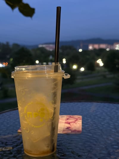 The image is a close-up, rotated shot of a plastic cup with a straw, sitting on a textured blue table. The cup appears to contain a beverage with ice and is slightly fogged from condensation. There is a pink and purple patterned napkin or wrapper behind the cup. In the background, out of focus, are lights from a city or buildings and trees, suggesting an outdoor setting at dusk or night. The overall mood is relaxed and casual.

Visible text on the cup, in yellow lettering, appears to be in Cyrillic script and includes the word "ПРО" (PRO). There are also circular bubble-like graphics on the cup.

The image was taken in Vladikavkaz, Russia. The time of day is likely evening or night, given the presence of city lights and the dim ambient light. The weather is not explicitly indicated, but the condensation on the cup might suggest it is warm, or the drink is cold.