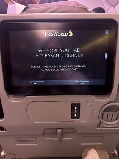 The image depicts the interior of an aircraft cabin, specifically a passenger's view of the seat-back in front. The primary subject is an illuminated in-flight entertainment (IFE) screen displaying the "KRISWORLD" logo and the Singapore Airlines emblem. The screen shows a message: "WE HOPE YOU HAD A PLEASANT JOURNEY" and "PLEASE TAKE YOUR BELONGINGS WITH YOU AS YOU LEAVE THE AIRCRAFT." Below this, "Local Time at Singapore 2:01 pm" is displayed on the left, and "Local Time at Brussels 7:01 am" is displayed on the right. An 'X' button for closing is visible in the upper right corner of the screen.

Below the screen, a passenger control panel is visible, featuring three vertical buttons: a call button (person icon), a fasten seatbelt/no smoking light (seatbelt icon), and a reading light button (light icon), which is illuminated. To the right of the control panel, a small storage pocket is integrated into the seat-back. A hinged tray table latch is visible below, and further down, a small white sticker containing safety instructions ("Fasten seat belt while seated" and "Life vest under your seat") is partially visible. On the far left, a headphone jack and a USB port are partially obscured.

A passenger's foot, clad in a light-colored sock or slipper, is visible in the bottom right corner of the frame. The upper portion of another passenger's head or seat is visible in the background, out of focus. The scene indicates the conclusion of an international flight arriving at Brussels Airport, which is located in Steenokkerzeel, Belgium.