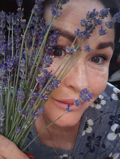 A close-up, vertically oriented, medium shot captures a woman's face as she holds a bouquet of lavender flowers. The flowers are a vibrant purple and green, arranged in a way that they partially obscure her face. Her skin is fair, with visible wrinkles around her eyes, suggesting she is middle-aged or older. Her eyes are a striking green, with dark lashes and well-defined eyebrows. Her lips are slightly parted, revealing a hint of her teeth. A thin, gold-colored chain is visible around her neck, resting on her décolletage, which is adorned with a grey blouse featuring a floral pattern of white and dark blue flowers. The background is dark and out of focus, making the woman and the lavender the clear subjects of the image. The lighting is soft, suggesting an indoor setting or perhaps a cloudy day. The overall mood is peaceful and serene, with the lavender adding a touch of natural beauty and perhaps a sense of calm. The woman's expression is neutral, but the way she holds the lavender suggests a gentle interaction with the flowers. There is no visible text in the image. The location context is Nova Kakhovka, Ukraine.