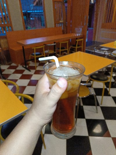 The image captures a first-person perspective shot within a casual eatery or cafe, likely in Ciamis, Indonesia. The primary subject is a person's hand holding a tall glass of iced tea, complete with ice cubes and a white bent straw. The hand appears to be light-skinned and slightly hairy.

The setting is an indoor space featuring a distinctive black and white checkerboard tiled floor. The furniture consists of bright yellow square tables and an orange booth-style bench seating arrangement along one wall, complemented by individual yellow and black metal chairs with round seats. Some tables appear to have metal legs. In the background, what looks like a window or doorway shows a blurred, dark blue or purple hue, suggesting it might be evening or night outside. The interior is artificially lit, contributing to the warm, somewhat grainy aesthetic of the photo.

No specific activity beyond holding the drink is depicted. There is no visible text in the image. The overall atmosphere appears relaxed and informal.