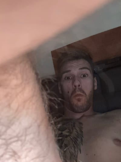 The media file shows a close-up, vertically oriented image of a person's face. The individual appears to be male, with light brown hair, brown eyes, and a beard. He has a mole on his chin and what looks like a small mark or blemish above his upper lip. The lighting is dim, suggesting an indoor setting, possibly in the evening or with low ambient light. The background is out of focus, showing dark and lighter brown shapes that could be furniture or a wall. A portion of what appears to be the person's chest or shoulder is visible in the upper right corner. A fuzzy, dark, spiky object is partially visible to the right of the person's head. The person's expression is neutral, with eyes looking directly at the camera. There is no visible text in the image. The overall impression is of a casual, candid shot.