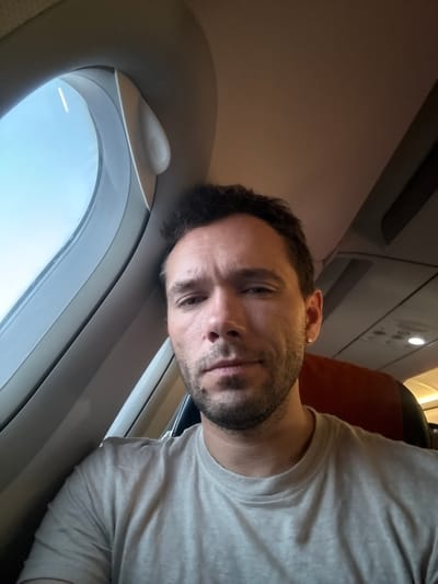 A man is taking a selfie in an airplane cabin, seated next to a window. The man is wearing a light-colored t-shirt and has short, dark hair and stubble. He has a small, skull-shaped earring in his left ear. The window shows a clear blue sky. The interior of the airplane is visible in the background, including seats and overhead compartments. The man appears to be looking at the camera. The image does not contain any information related to the city of Abakan, Russia.