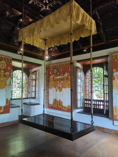 An antique wooden swing, known as an *oonjal*, dominates the center of a well-lit room in the Mattancherry Palace, Kochi, India. The swing features a rectangular, dark brown, polished wooden platform, suspended by four black metal chains. Each chain attaches to a light-colored wooden pole, which extends upwards to support a rectangular, ruffled, golden fabric canopy. A small, dark brown sign with "DON'T TOUCH" in white text rests on the swing's platform.
The room's walls are light-colored, serving as a backdrop for three large traditional Kerala mural paintings. The central mural, on a vibrant red background, depicts multiple figures in traditional attire, possibly deities or royal figures, one holding a musical instrument. Portions of two additional murals, each showing a standing figure in white, are visible on the left and right walls.
The dark wooden ceiling above is ornately carved with intricate patterns, and several recessed light fixtures illuminate the space. Three multi-paned wooden windows are visible along the right wall, with one on the left. The central right window features an intricate dark metal grille, and glimpses of green foliage are visible through the open windows, suggesting a garden exterior. The floor is made of polished wooden planks.