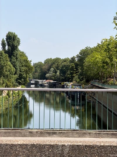 The image shows a view of the Seine River in Neuilly-sur-Seine, France, from a bridge. The river is lined with lush green trees on both sides. A row of houseboats is moored along the riverbank, some with people visible on deck. The water reflects the trees and the sky. The foreground shows the railing of the bridge and a concrete pathway. The sky is clear and blue.