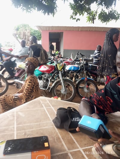 In Dokan Tofa, Nigeria, a group of people are gathered around several motorcycles, suggesting a marketplace or transportation hub. A woman in a patterned brown and black dress and headscarf is seated in the foreground, looking towards the camera. To her right, another person's hands are visible holding a blue payment terminal. In the background, a woman in a black head covering with gold embellishments is standing near a motorcycle. Several other individuals and motorcycles are present, creating a scene of daily life and commerce. The environment appears to be outdoors, possibly under the shade of trees, with a building with a pink wall visible behind the subjects. The presence of a payment terminal indicates a transaction is taking place or has recently occurred.