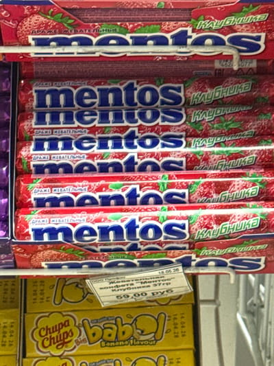 The image shows a close-up view of several rolls of Mentos strawberry-flavored chewy candies displayed in a store or market. The rolls are primarily red with blue lettering spelling out "mentos." Some of the rolls also have images of strawberries and green leaves on their packaging, along with text in Cyrillic. A small white price tag is attached to one of the rolls, displaying information in Cyrillic including a price of "59.00 руб" (59.00 rubles) and a date of "18.05.26". To the right of the Mentos rolls, there are yellow boxes of Chupa Chups candy, with "Chupa Chups" and "Banana Flavour" visible. Dates such as "14.04.26" and "14.04.28" are printed on the Chupa Chups boxes. The overall setting appears to be a retail display shelf. There are no people visible in the image. The lighting suggests an indoor environment, likely a store with artificial lighting.