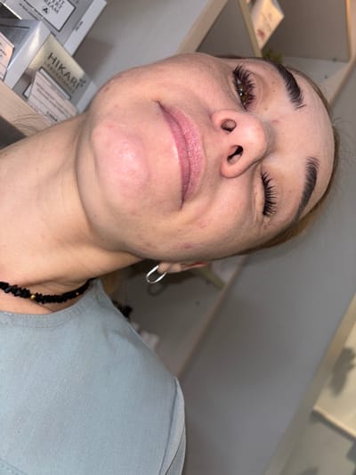 The image shows a close-up, slightly tilted, overhead view of a person's face and upper chest. The person appears to be female, with fair skin, dark eyebrows, and visible eyelashes. Her mouth is slightly open, revealing her tongue. She is wearing a light blue top and a black beaded necklace with gold accents. A silver hoop earring is visible on her right earlobe. In the background, shelves are visible with boxes of cosmetic products. The environment appears to be indoors, possibly a beauty salon or retail space. The specific location in Ladozhskoe Ozero, Russia cannot be determined from the image alone, as there are no identifying landmarks or context clues to suggest this location.