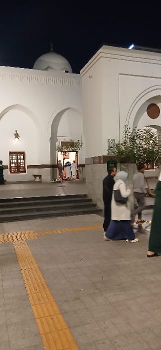 This nocturnal image depicts the exterior of the Quba Mosque in Medina, Saudi Arabia. The prominent building is constructed of white-painted material, featuring a large central dome topped with a gold finial, multiple arched facades, and a decorative fluted parapet. A main entrance, brightly illuminated, is visible at the center-left, accessed by a set of dark steps. Inside this archway, several individuals, some attired in traditional white thobes, are observed entering or exiting.

The ground in the foreground consists of light grey square paving tiles, marked with an L-shaped pattern of yellow tactile strips. Approximately five individuals are present on this paved area. In the center-right, a female figure wears a light-colored hijab, a long beige coat, and a dark blue skirt, carrying a black shoulder bag, positioned next to a figure in dark clothing. To their right, a male figure in a light grey long-sleeved top and dark trousers walks towards the left. A partially visible figure in a dark green garment is on the far right. On the far left, another figure in dark green is partially visible near the base of the mosque. Small trees or shrubs are planted adjacent to the building walls, and a wooden bench is positioned under an archway on the left side. Artificial light sources illuminate the building and ground.