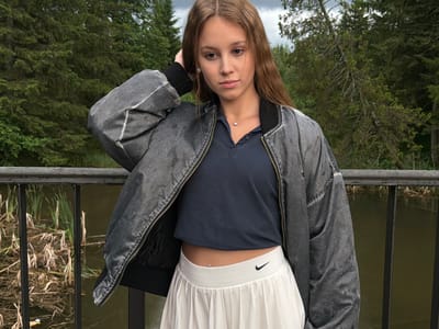 A young woman stands on a walkway behind a black metal railing, with a body of water and marshy vegetation to her left. She is wearing a dark blue polo shirt, a grey bomber jacket, and a white pleated skirt with a Nike logo. Her hair is long and brown, and she has a belly button piercing. The background consists of dense green trees. The image appears to have been taken outdoors during the day. The location is identified as Chusovoy, Russia.