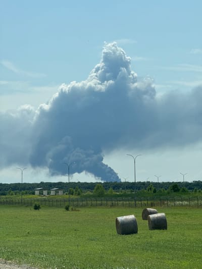 The image shows a wide, grassy field with three large hay bales in the foreground. A fence with barbed wire runs across the middle ground, separating the field from a line of trees and some industrial-looking structures with peaked roofs. Beyond the trees, a massive plume of dark smoke billows into a clear blue sky, obscuring a distant horizon. Several tall lampposts are visible along the tree line. The scene is devoid of any people. The location is identified as Rozhdestvenskaya, Russia.