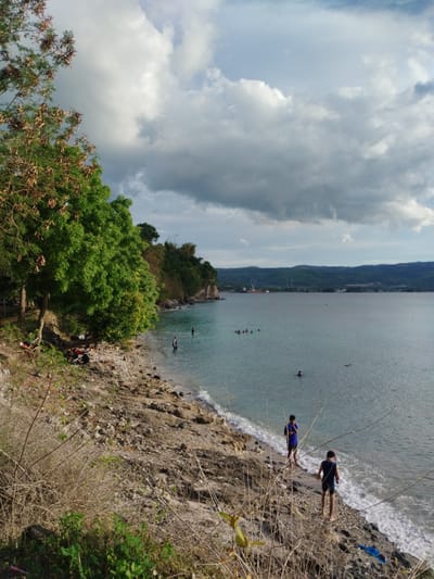 The scene is a coastal area in Lamreh, Indonesia, showing a rocky beach, ocean, and sky with clouds. Two boys stand near the water's edge, the closer one walking towards the water and the other standing still. Several people are swimming in the ocean. A tree with green and brown leaves is located on the left side of the frame. The foreground contains dry vegetation. In the distance, there is a coastline and land with visible industrial structures.