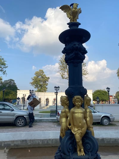 A person carries a stack of cardboard boxes on their head in front of a white building with arched windows. The person is walking on a paved area with a silver car to their left and another silver car to their right. In the foreground, a black fountain with gold cherub statues is visible. The sky is blue with scattered white clouds. The scene appears to be in Kathmandu Metropolitan City, Nepal.