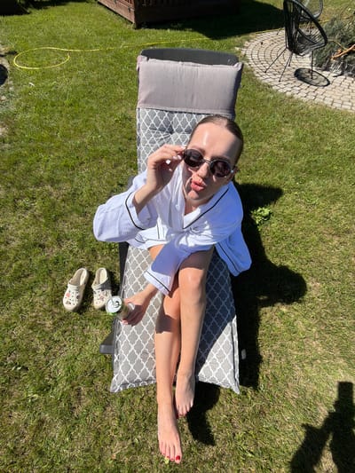 A woman is lying on a lounger on a sunny day in Dreiliņi, Latvia. She is wearing a white robe and sunglasses, and her legs are stretched out on the lounger. She is holding a can of drink in her right hand. Next to the lounger are a pair of Crocs shoes. The scene is set in a grassy backyard with a paved area and some outdoor furniture in the background. The sun is casting long shadows, indicating it is likely late afternoon. The woman appears relaxed and is enjoying the warm weather.