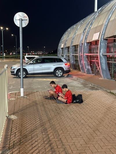 This night-time image, taken in Lod, Israel, shows an urban scene featuring two young males, a silver car, and distinctive modern architecture under artificial illumination.

On the right side of the frame, two young males are squatting or sitting on a reddish-brown brick sidewalk. Both are wearing red short-sleeved shirts and dark shorts, and are deeply engrossed in their mobile phones. One of them, closer to the viewer, has a dark backpack resting beside him.

To their left, a silver SUV or crossover vehicle is parked or stopped by the curb on an asphalt road. Its taillights are illuminated. Further to the left and extending upwards into the frame is a prominent, multi-story, curved structure made of glass and metal, possibly a covered walkway or part of a building. This structure features red accent lighting within its framework. The dark road stretches into the background, lined with streetlights and showing the distant lights of other vehicles. In the lower left foreground, a metal pole with the unreadable back of a circular sign partially obstructs the view of the road and the car.

The overall scene depicts a casual moment in a well-lit urban environment at night, with the individuals focused on their digital devices. There is no visible text identifiable in the image.