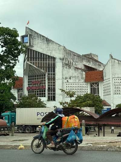 A person rides a motorcycle loaded with goods in front of a building with Vietnamese writing. The building is white and has a distinctive angular design. A large clock is visible on the upper left side of the building, and a flag is planted on the peak. The writing on the building reads "CHỢ ĐÔNG HÀ", which translates to Dong Ha Market. A large truck with "POST" written on its side is parked behind the motorcycle. The ground is paved, and there are some trees and awnings visible in the background. The sky is overcast.