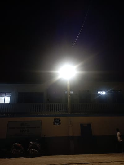 The image, captured at night in Dayahtuha, Indonesia, depicts a multi-story building illuminated by artificial light sources. A powerful street light or floodlight, creating a bright starburst effect and lens flare, is the dominant light source, situated near the center of the frame and brightly illuminating parts of the building. The building itself has a light-colored facade with visible windows, some featuring decorative grilles, and balcony railings. Lights are visible through several of the building's windows.
On the ground level next to the building, two motorcycles are parked. A person wearing a light-colored shirt and dark pants is partially visible on an upper level or balcony of the building. The ground to the right of the building appears to be an unpaved path.
The dark night sky, occupying the left side of the frame, shows a faint, diagonal line, possibly a distant light trail or aircraft contrail. A sign affixed to the ground floor of the building is partially visible, bearing the text "BALAI PELATIHAN PEMENUHAN GIZI" and the prominent acronym "SPPG". This translates to "Nutrition Fulfillment Training Center," indicating the building's function. The overall scene portrays a quiet, artificially lit night environment.